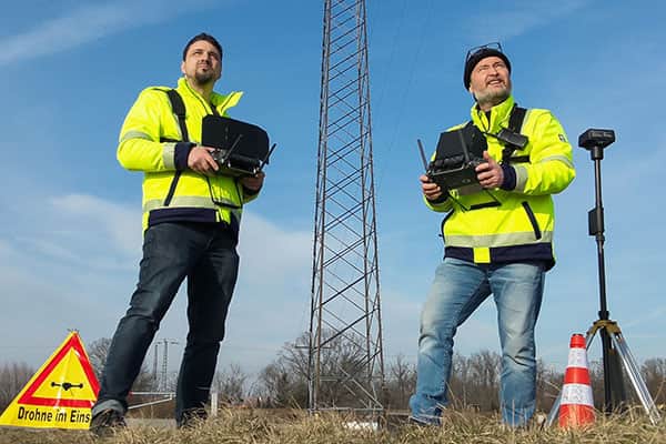 Zwei Piloten von Multikopterdienst steuern Drohne & Kamera, Sicherheitsmaßnahmen im Vordergrund, Strommast im Hintergrund